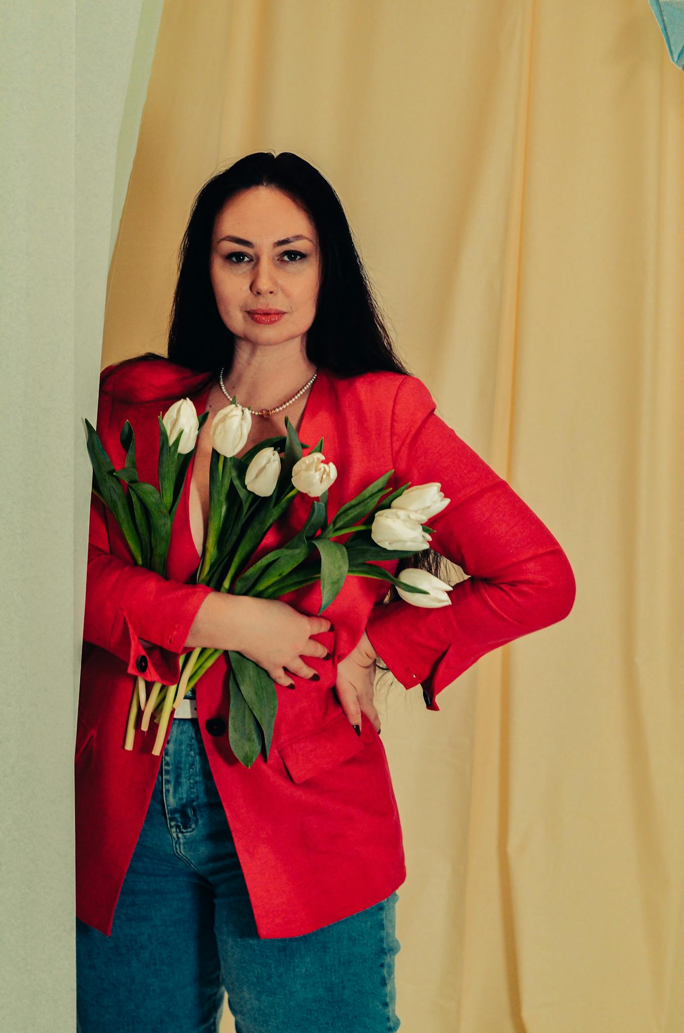 Elegant woman in a red blazer holding white tulips, posing with confidence against a neutral background.
