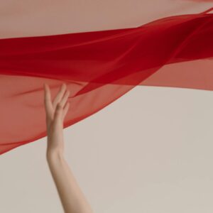 A delicate hand interacts with vibrant red fabric against a neutral background, creating artistic fluidity.