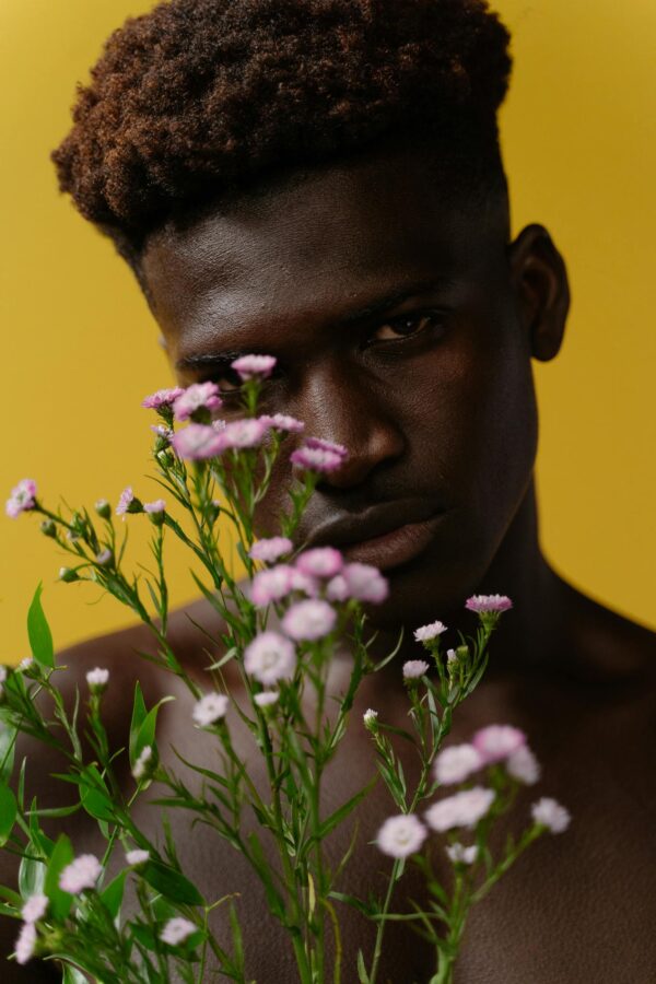Close-up portrait of a pensive man holding delicate pink flowers against a vibrant yellow background.
