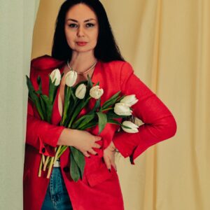 Elegant woman in a red blazer holding white tulips, posing with confidence against a neutral background.